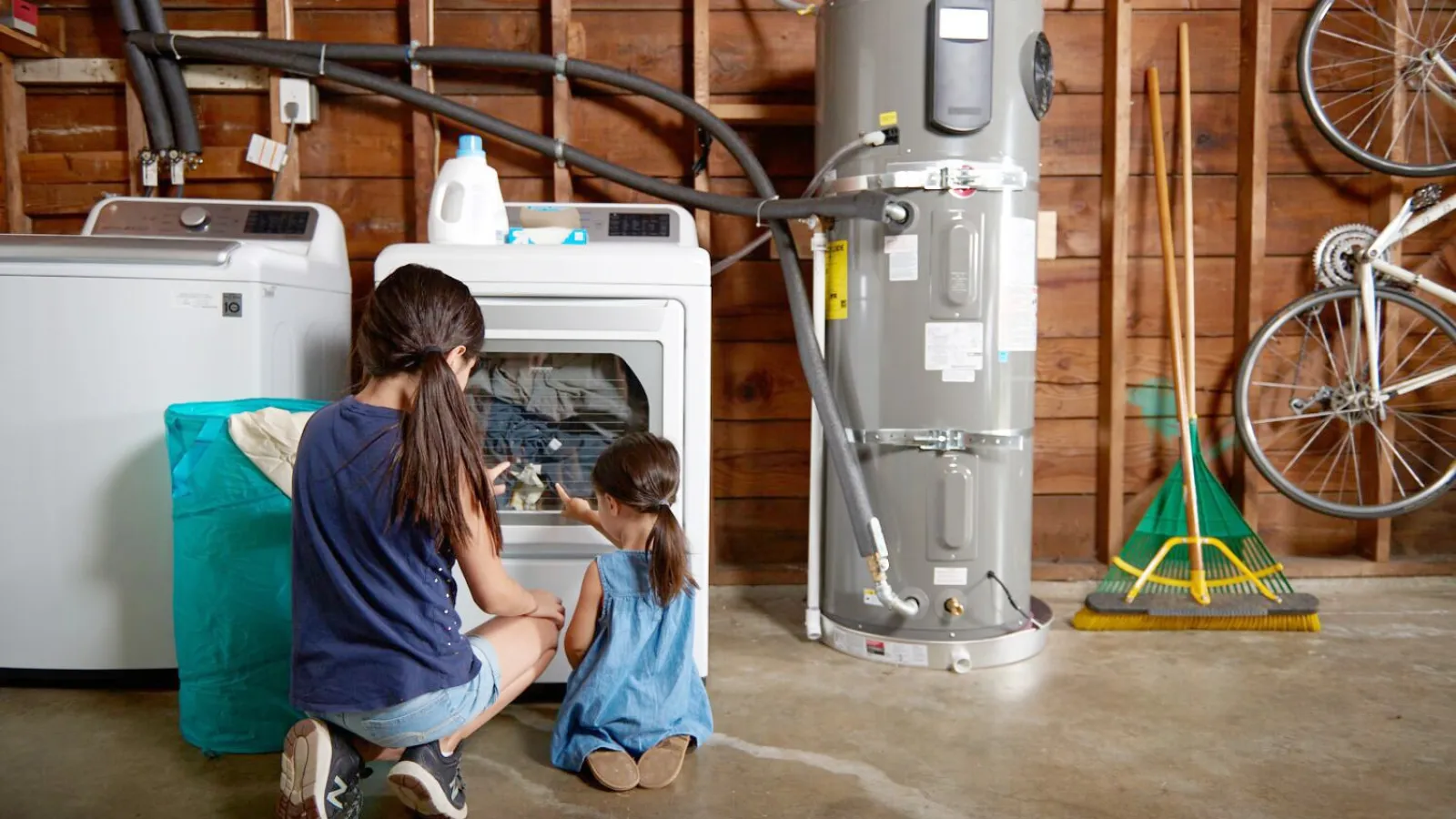 Two girls watching clothes inside a dryer in a home laundry room with water heater and tools nearby