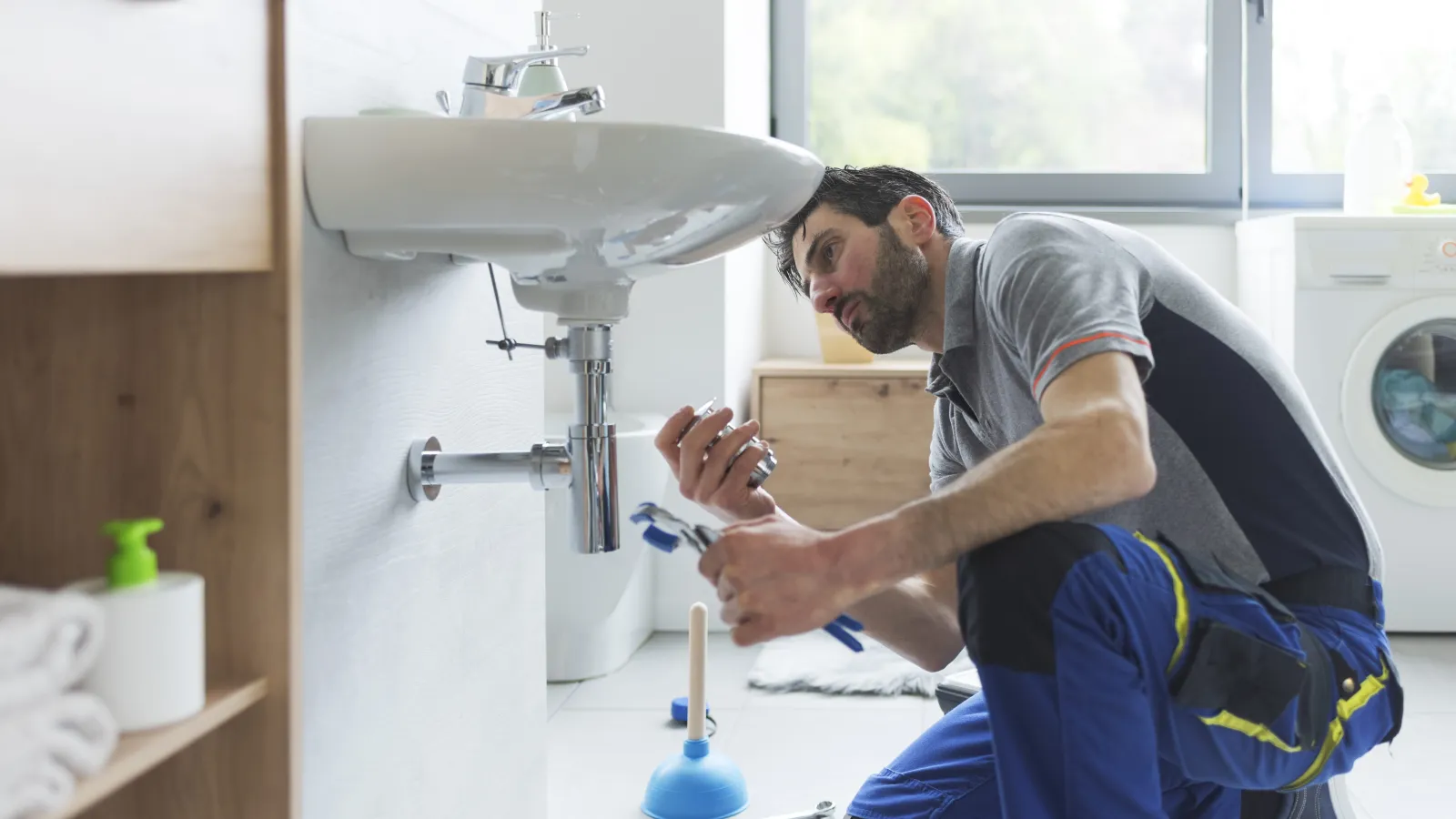 Plumber repairing a bathroom sink drain pipe with tools and plunger nearby in a modern home.