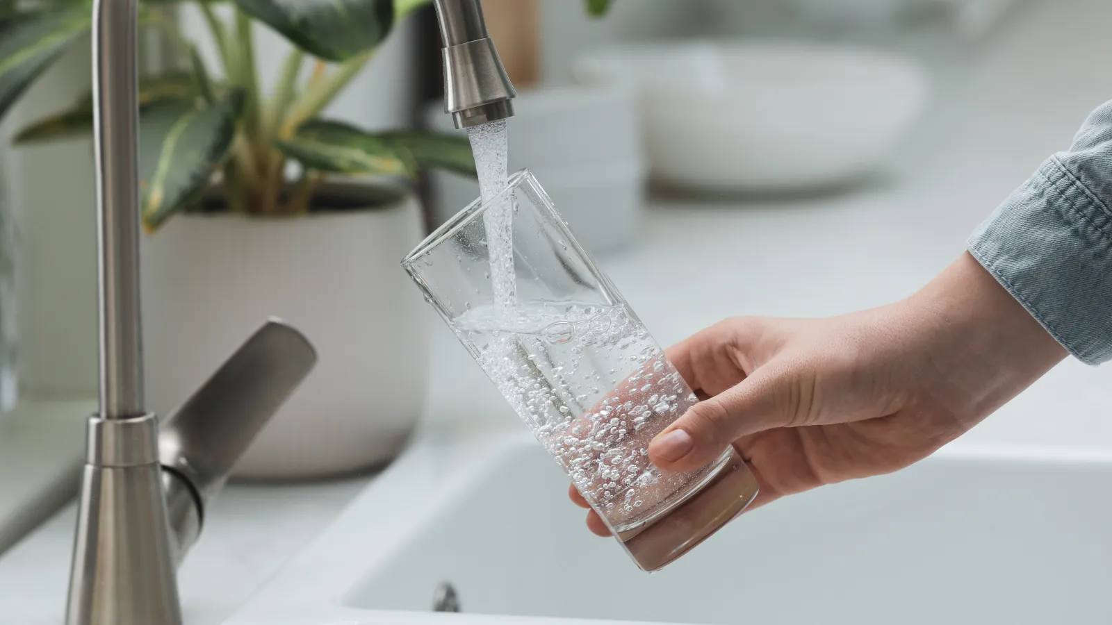 Hand holding a glass under kitchen faucet filling it with clear water in a bright setting with plants.