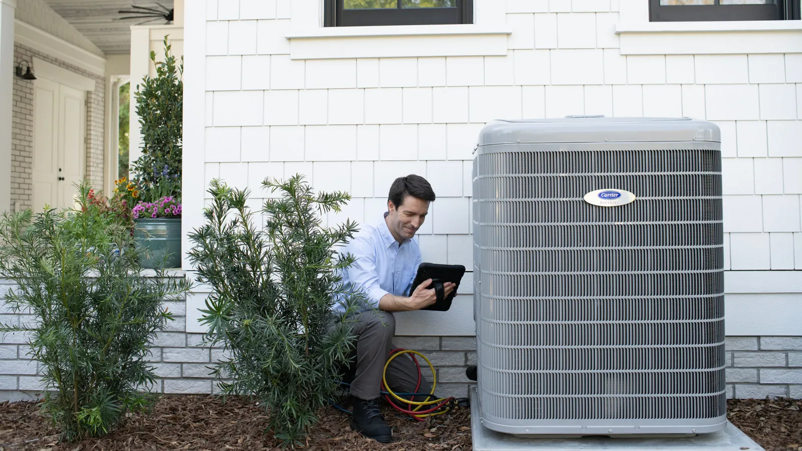 Technician inspects residential air conditioning unit beside house using a tablet with connected gauges.