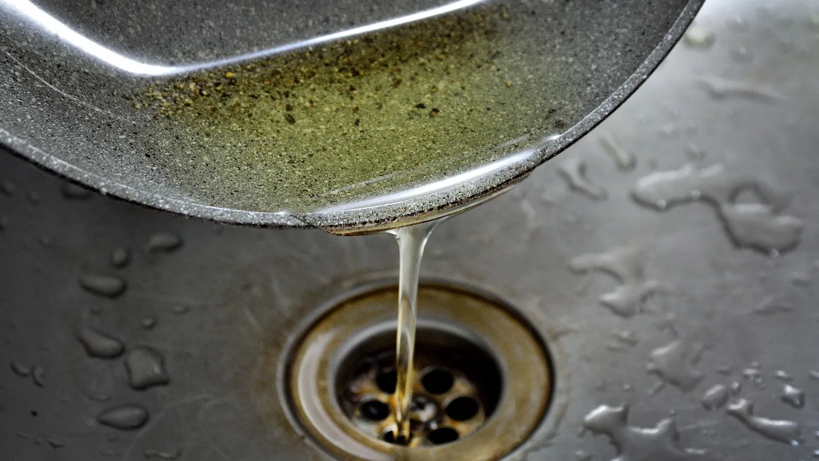 Used cooking oil being poured from a pan into a metal kitchen sink drain with water droplets nearby.