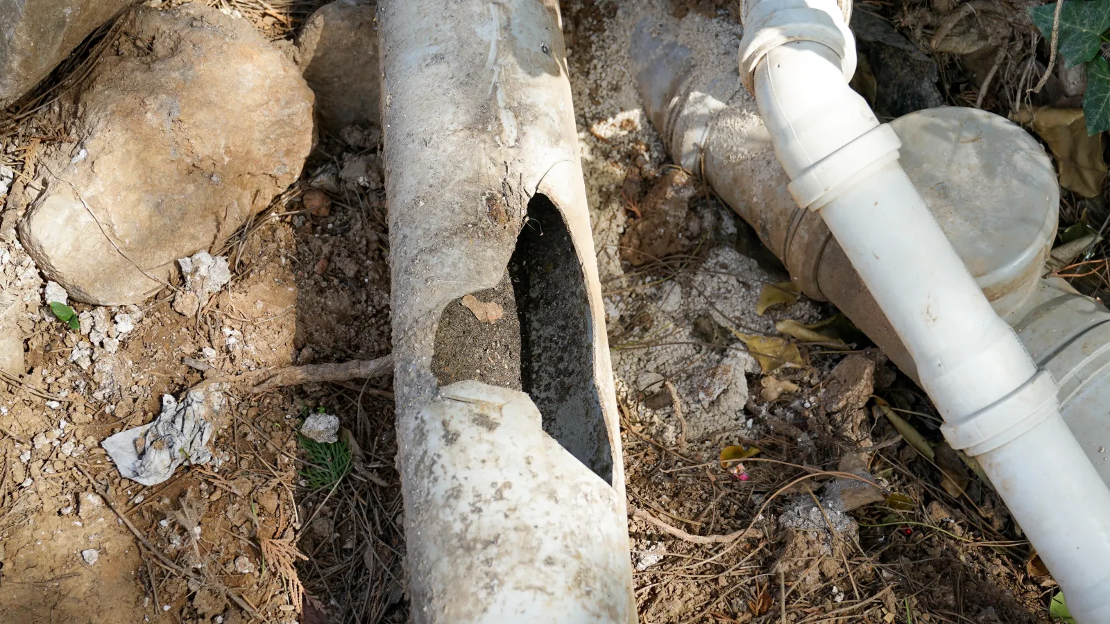 Old broken concrete pipe with a large hole next to intact white PVC pipes on dry ground with rocks.