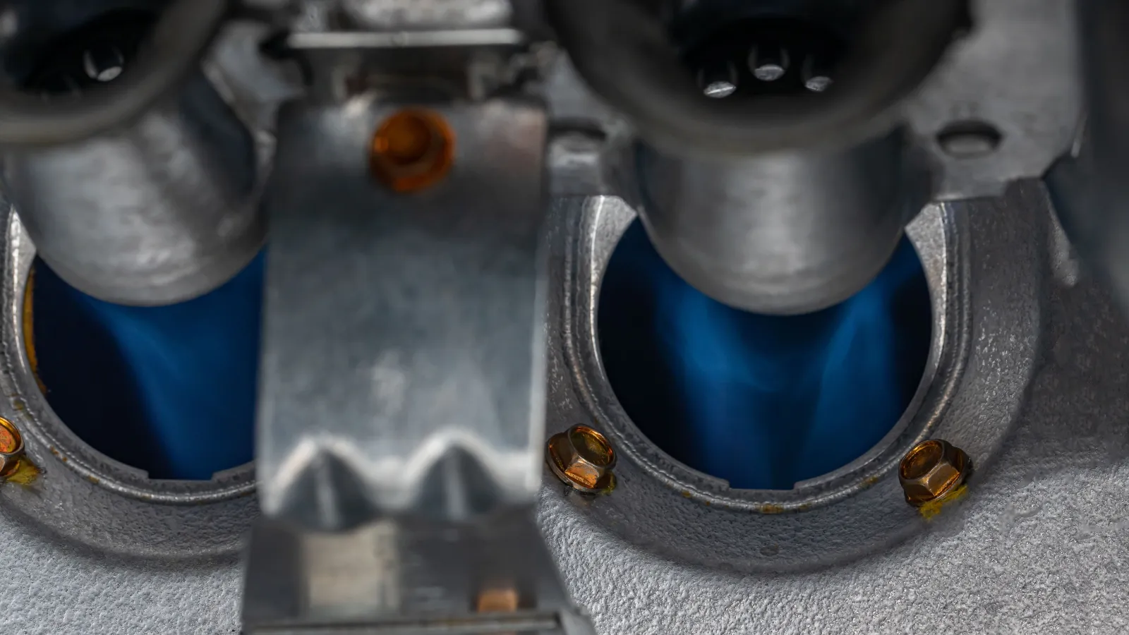 Close-up of blue gas flames burning inside a metal stove burner system with bolts and metallic parts.