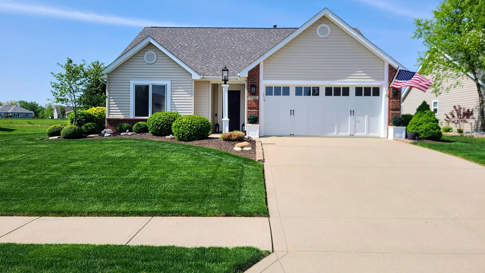 Single-story beige house with white garage door, well-maintained lawn, shrubs, and an American flag on the porch