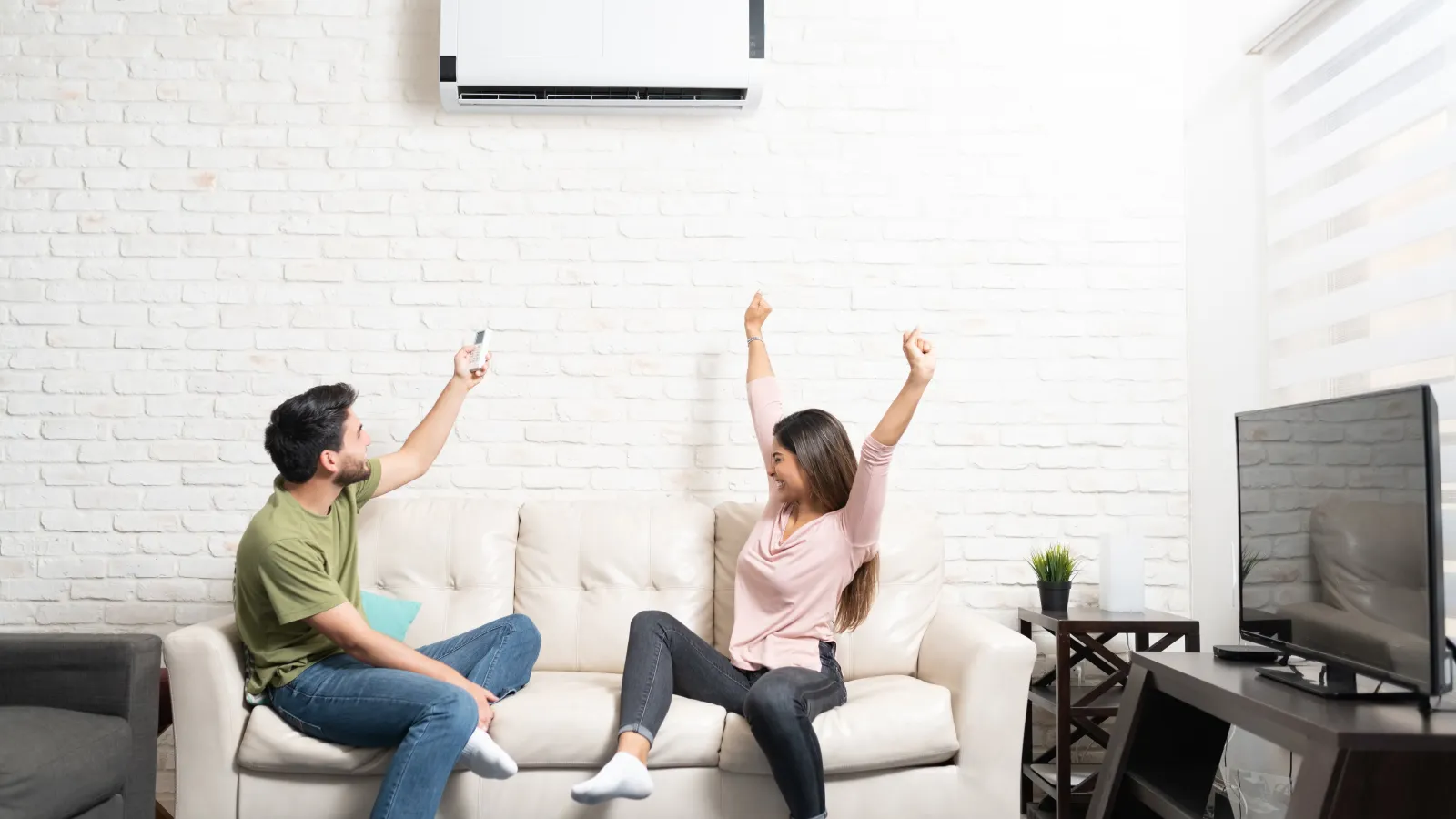 Happy couple sitting on sofa controlling air conditioner with remote in modern living room