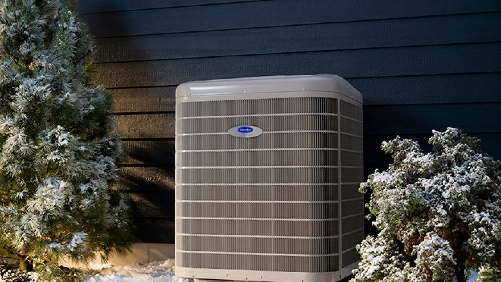 Outdoor air conditioning unit next to snow-covered evergreen trees against a dark blue house wall.