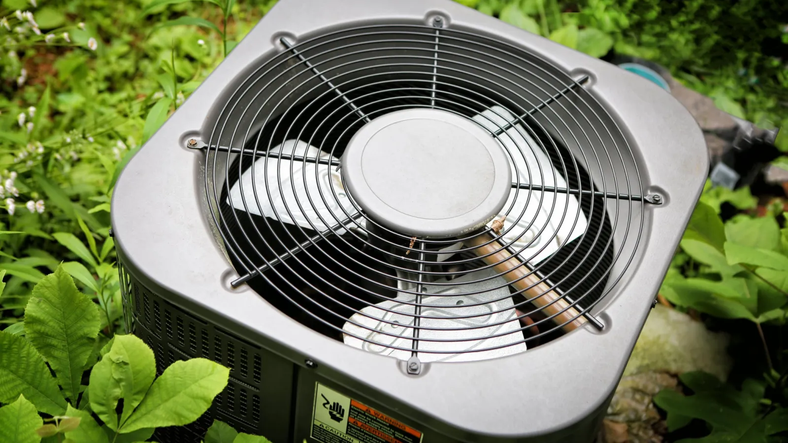 Outdoor air conditioning unit surrounded by green plants in a garden setting with metal fan cover.
