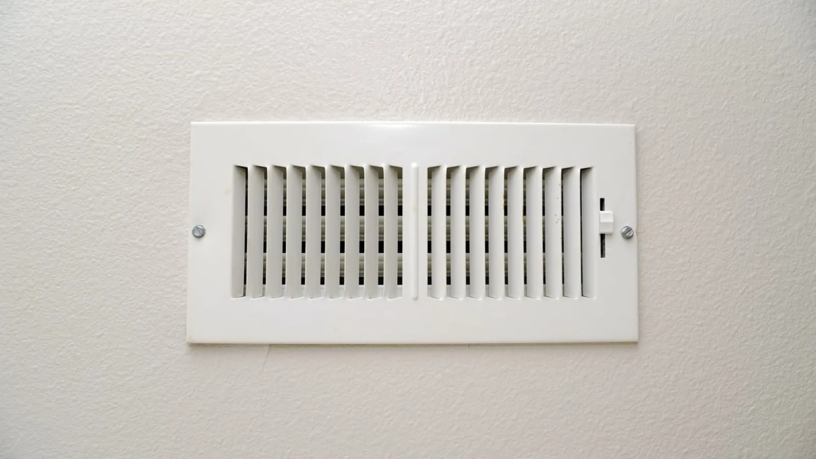 White rectangular air vent grille mounted on a beige textured wall with adjustable slats and screws.
