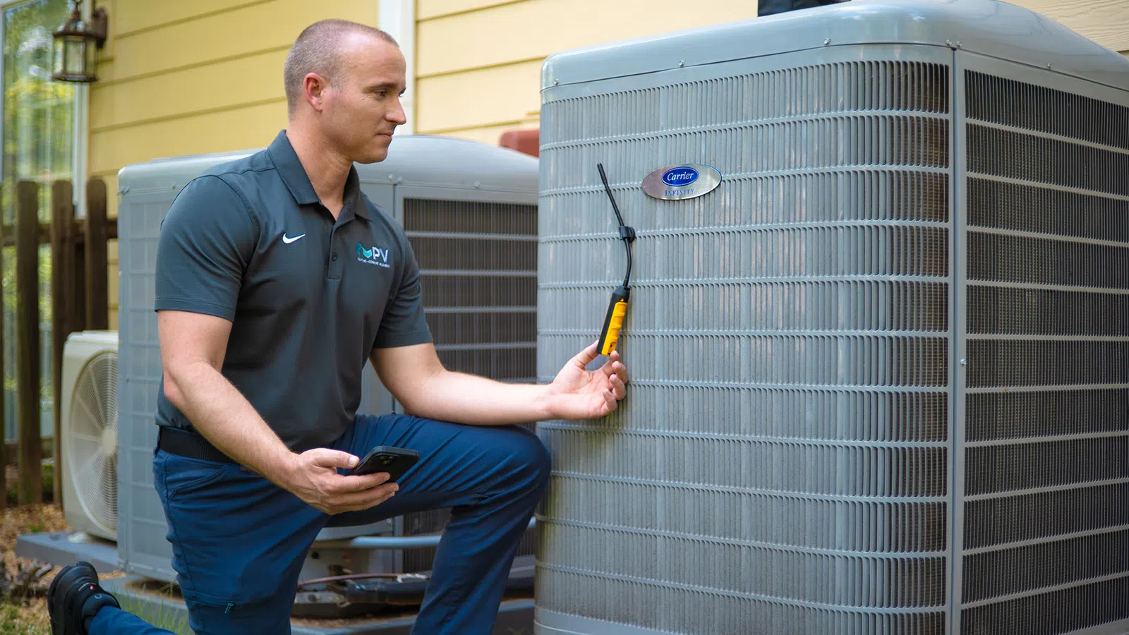 Technician inspecting an outdoor Carrier air conditioning unit using a diagnostic tool and smartphone in daylight.