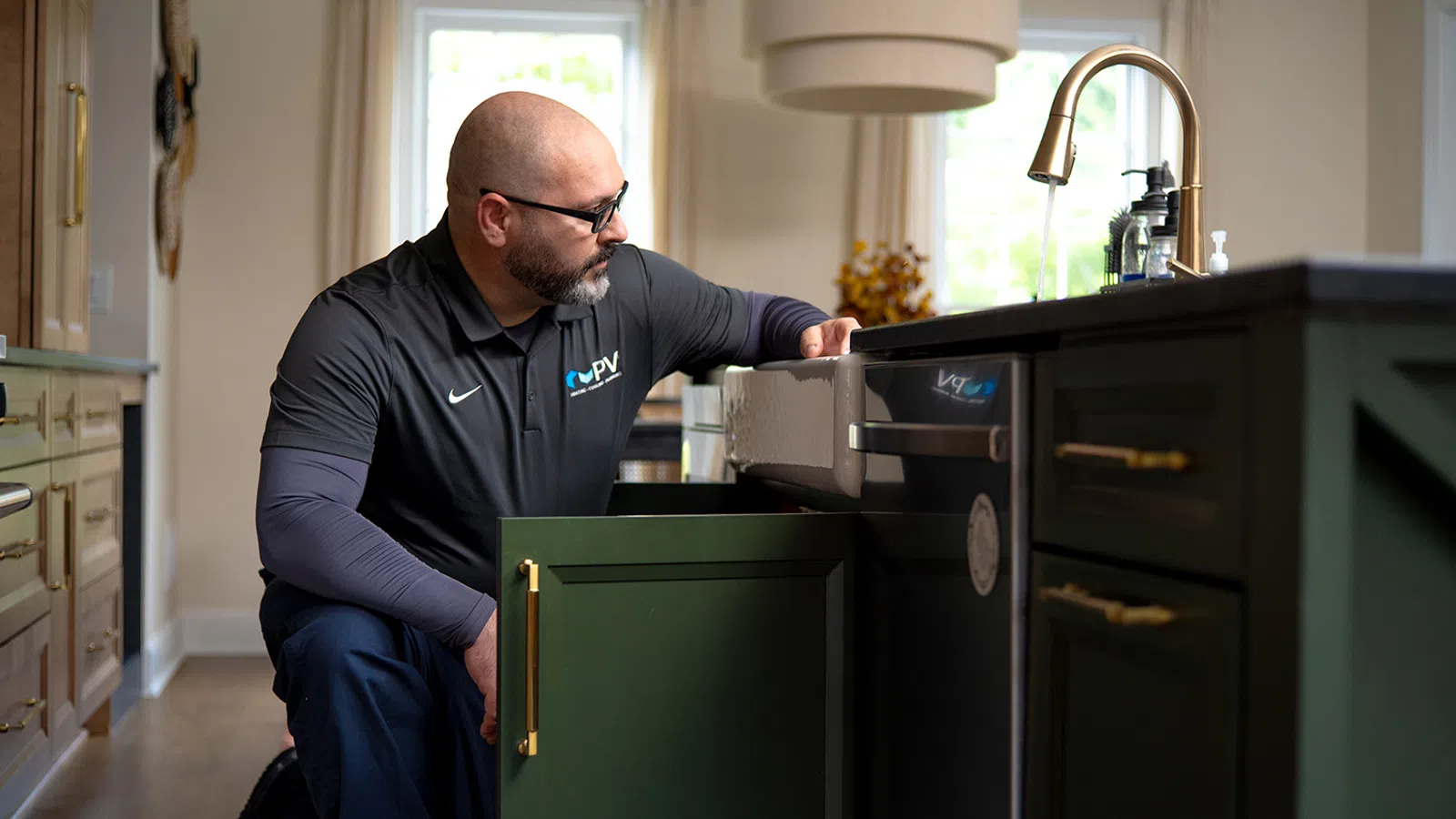 Technician inspecting plumbing under kitchen sink with green cabinets and gold handles in a modern home.