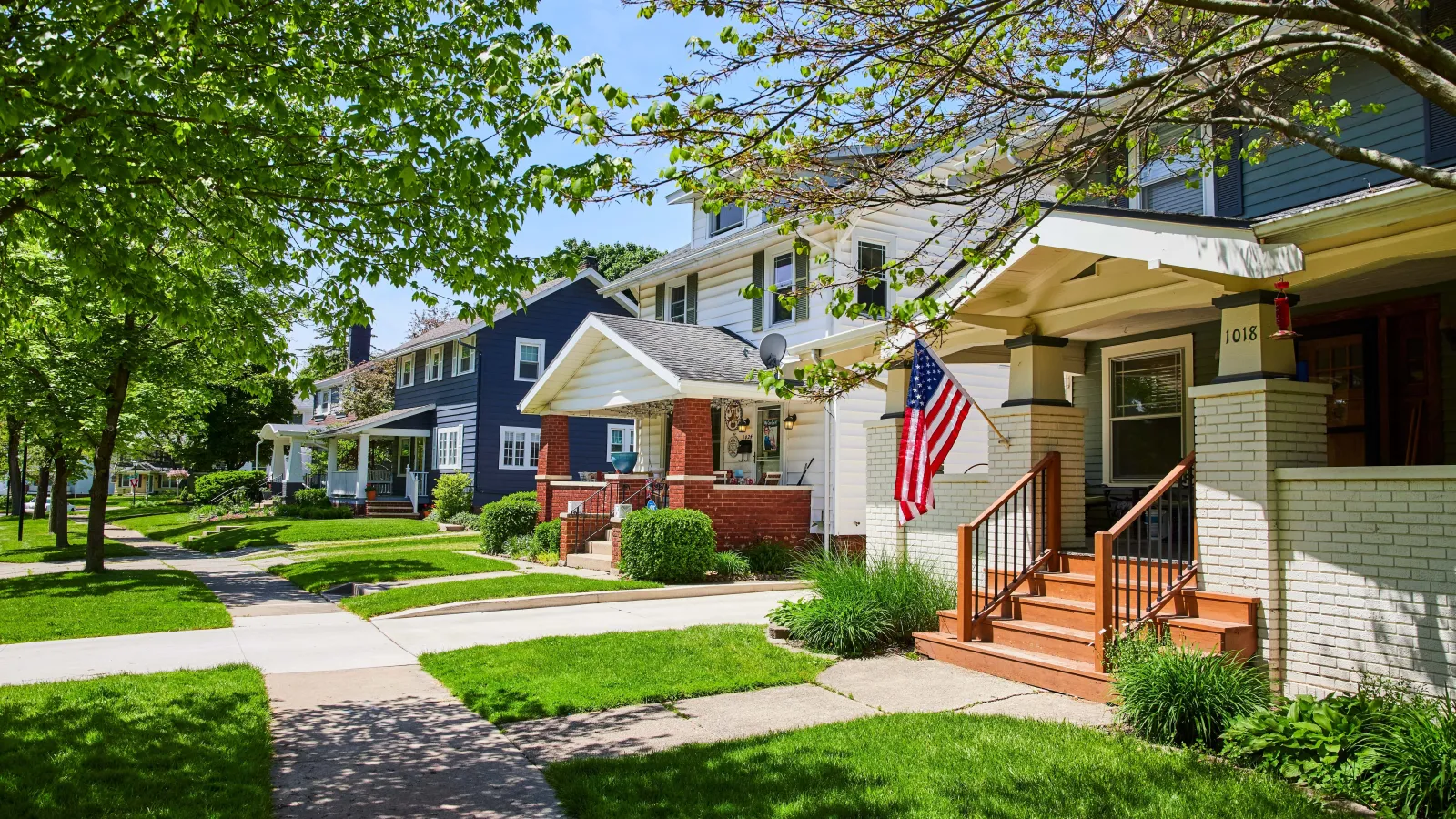 Tree-lined neighborhood street with American flag and colorful houses on a sunny day