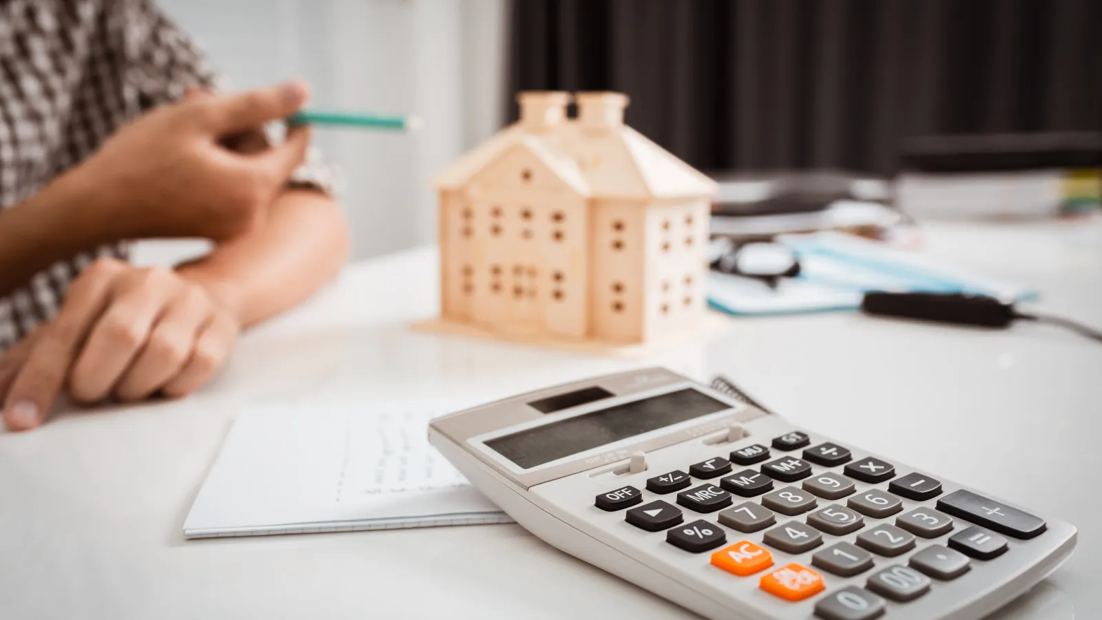 Calculator, notebook, and wooden house model on desk with person pointing, symbolizing home finance planning.