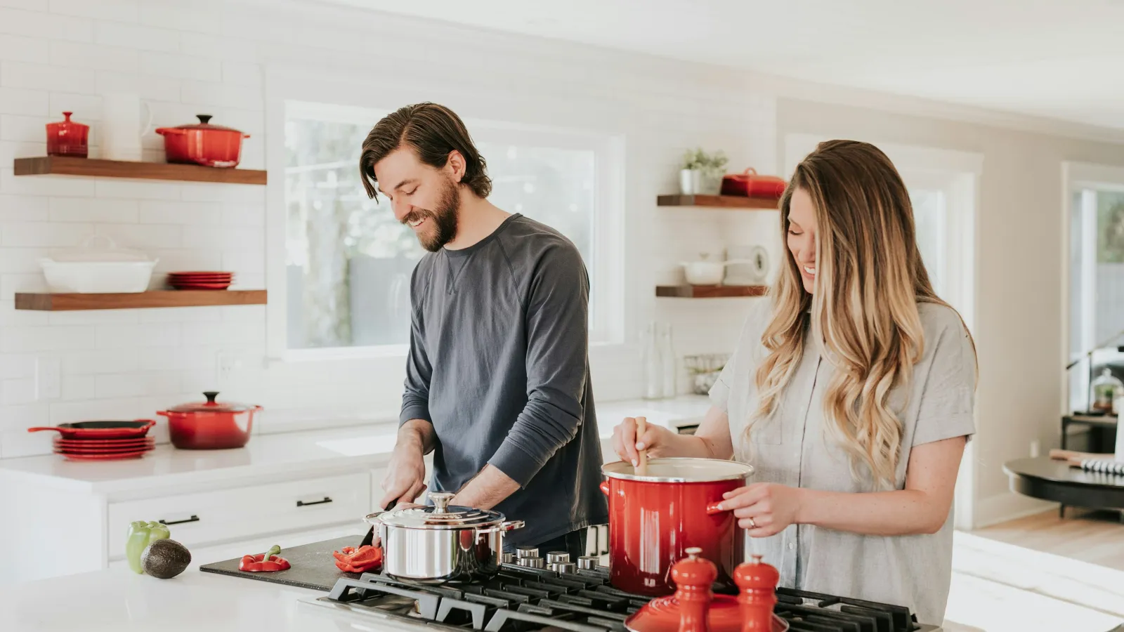 Couple cooking together in a bright kitchen with red cookware and fresh vegetables on the counter.