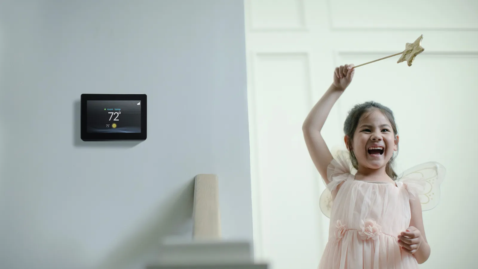 Smiling girl in fairy costume with wand next to wall thermostat set at 72 degrees in bright home hallway