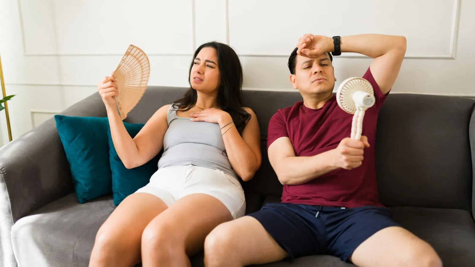 Man and woman sitting on couch in summer clothes using handheld fans to cool down indoors.
