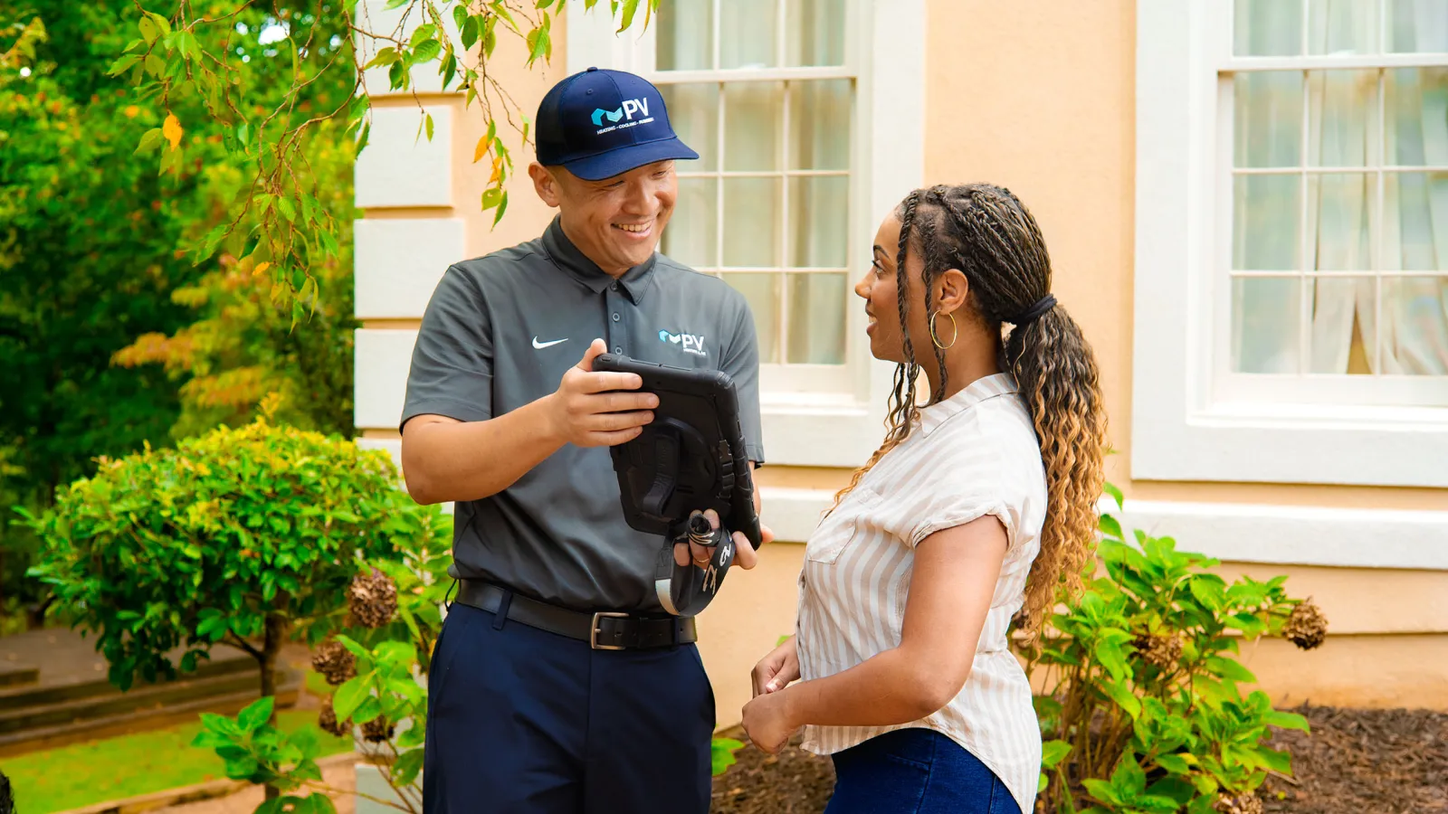 Technician in uniform showing a tablet to a woman outside a house with greenery and windows in the background