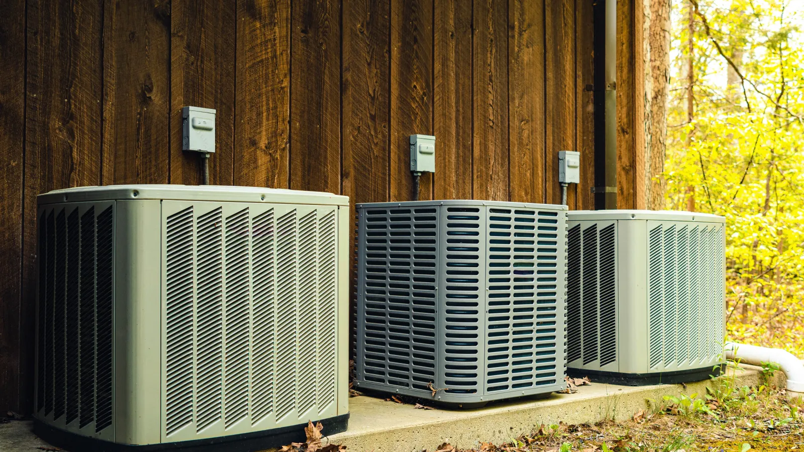 Three outdoor air conditioning units installed against a wooden wall surrounded by fallen leaves and greenery.