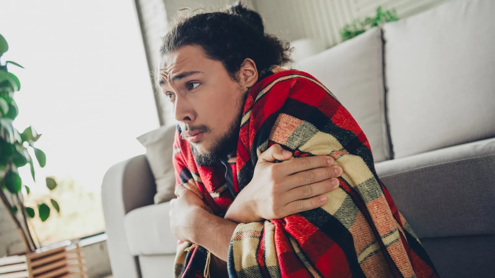 Young man wrapped in a plaid blanket sitting on floor shivering indoors near a gray sofa and window.