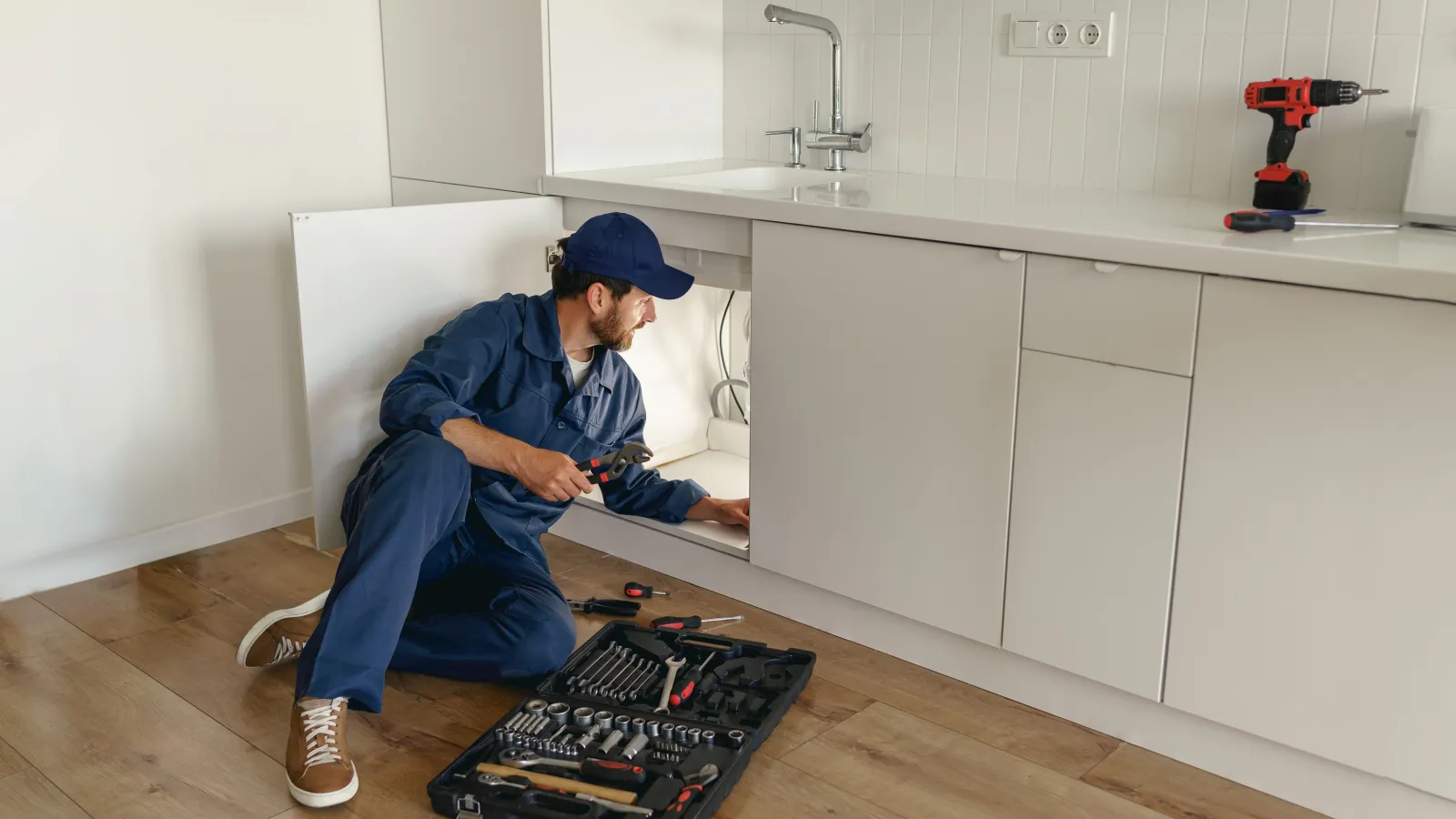 Plumber in blue uniform repairing pipes under kitchen sink with tools on wooden floor and cordless drill on counter