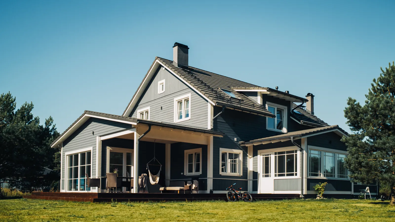 Modern gray two-story house with large windows, a covered patio, and a green lawn under clear blue sky.