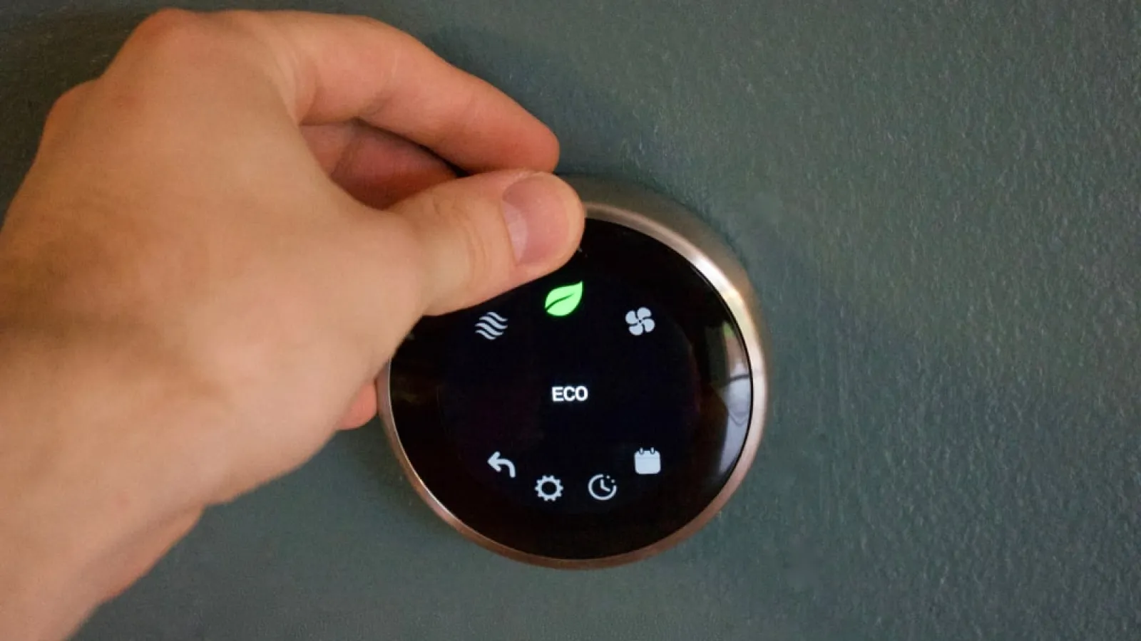 Hand adjusting a round digital thermostat set to eco mode on a dark teal wall.