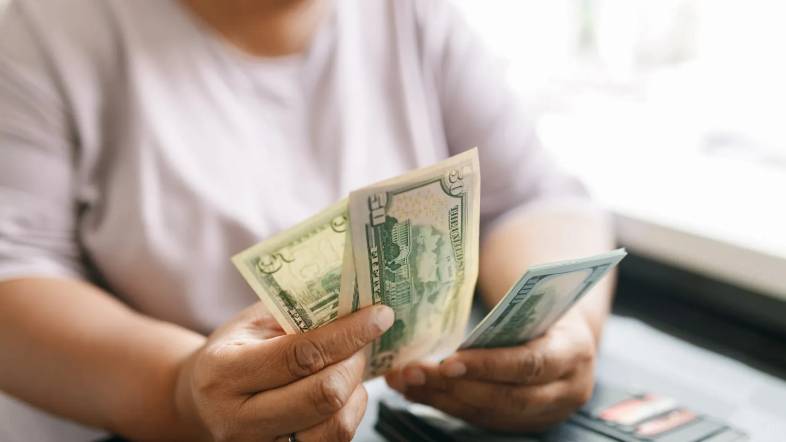 Person counting US dollar bills with a wallet on the table in natural light.