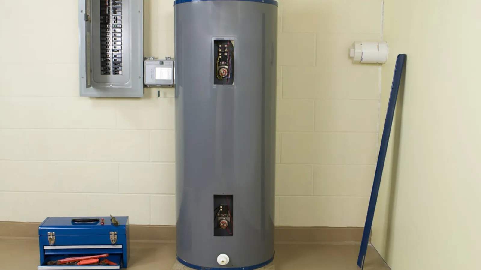 Gray water heater tank next to blue toolbox and electrical panel in a utility room with cream walls