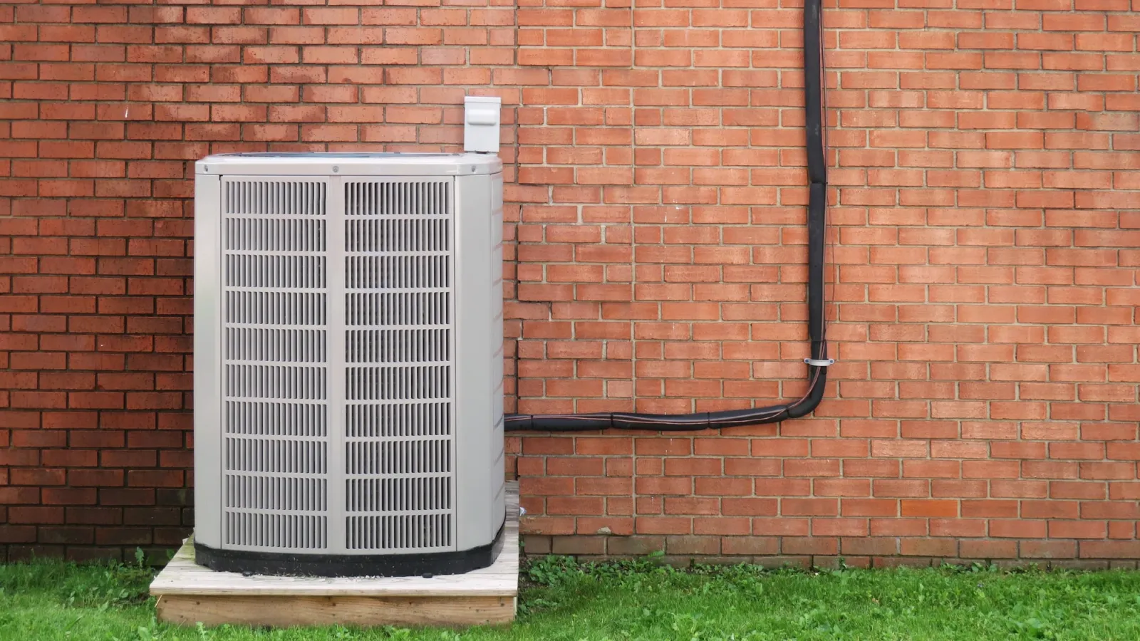 Outdoor air conditioning unit installed on wooden platform beside red brick wall with black conduit piping.