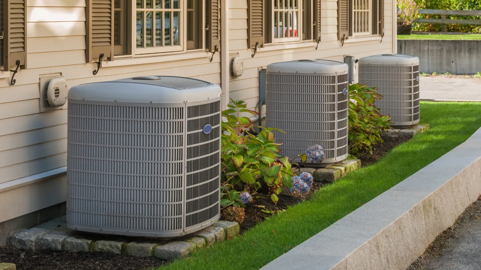 Three outdoor air conditioning units installed beside a beige house with green plants and stone foundation.