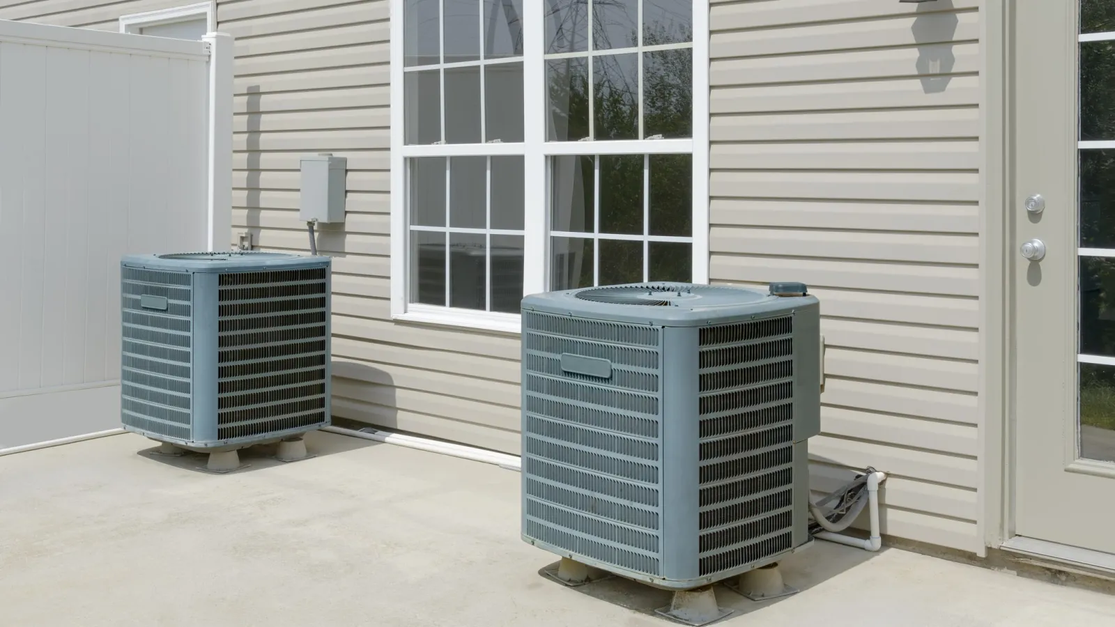 Two gray outdoor air conditioning units installed on concrete slabs by a beige house with a window and door.