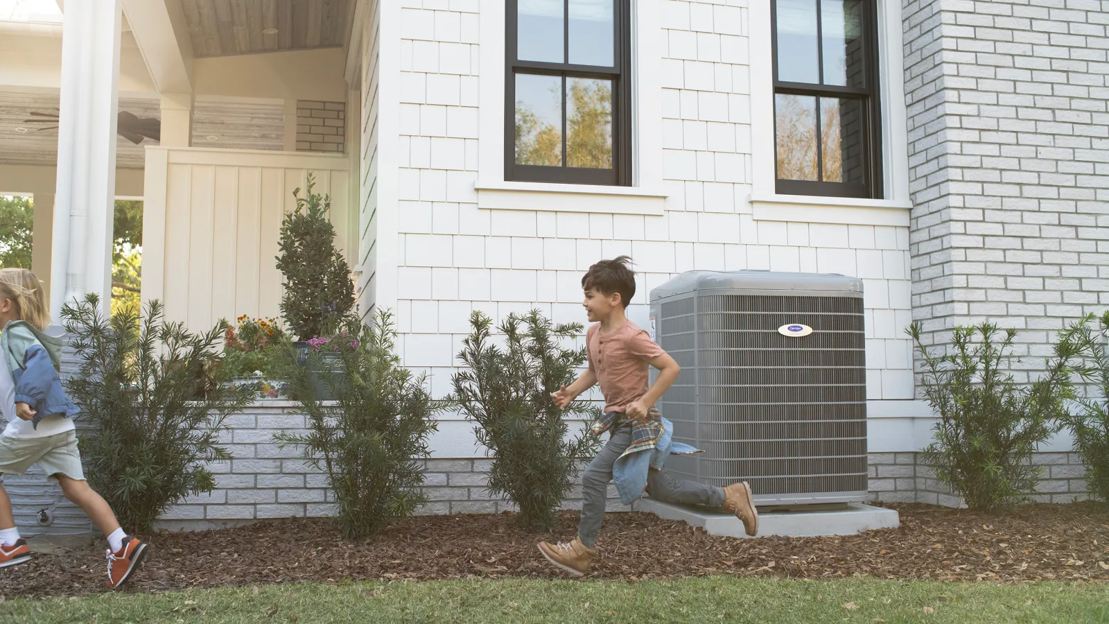Two children running and playing outside a white house with an air conditioning unit against the wall