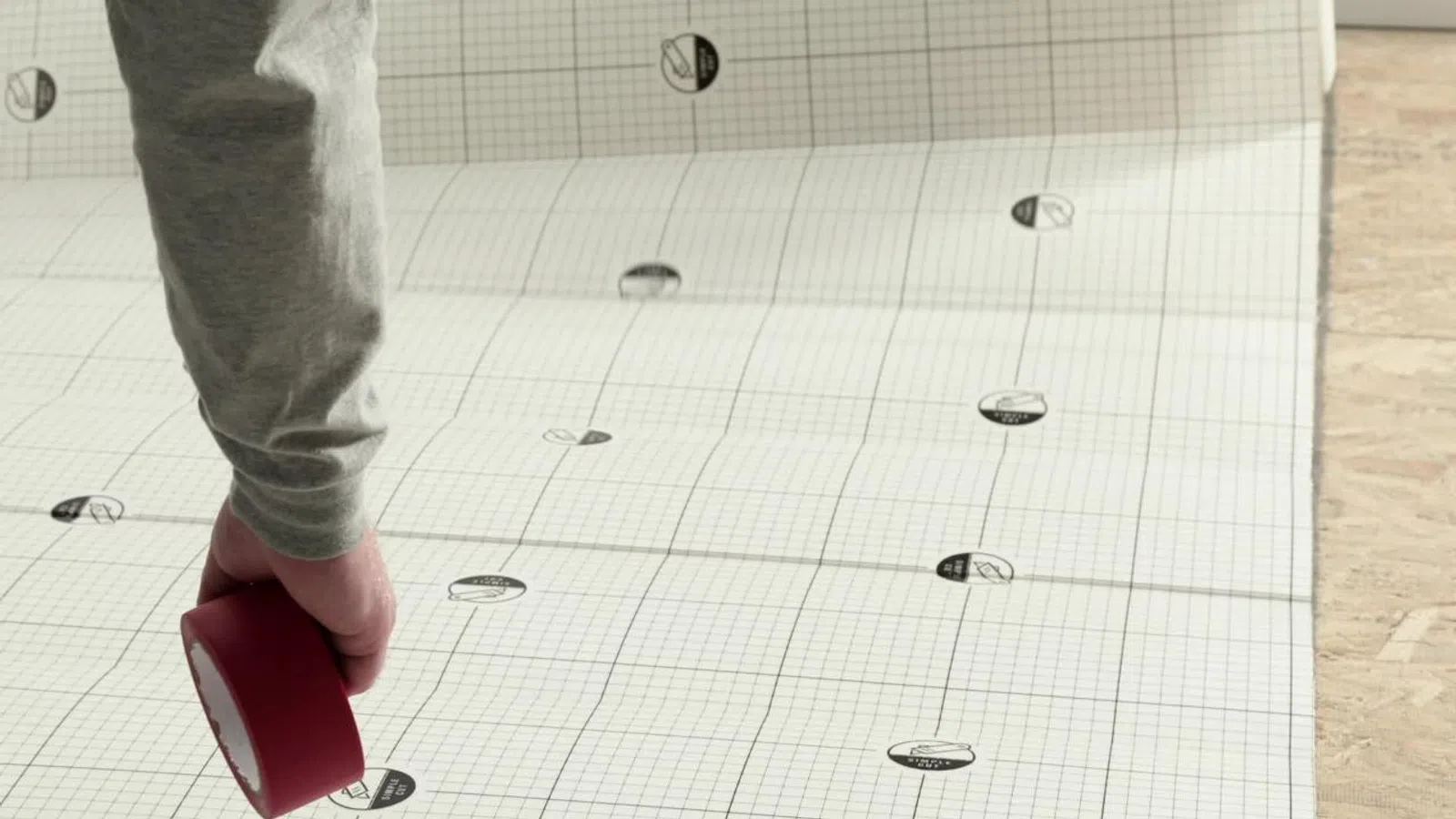 Person's hand holding red tape over grid-patterned paper with logos spread around on a wooden surface.