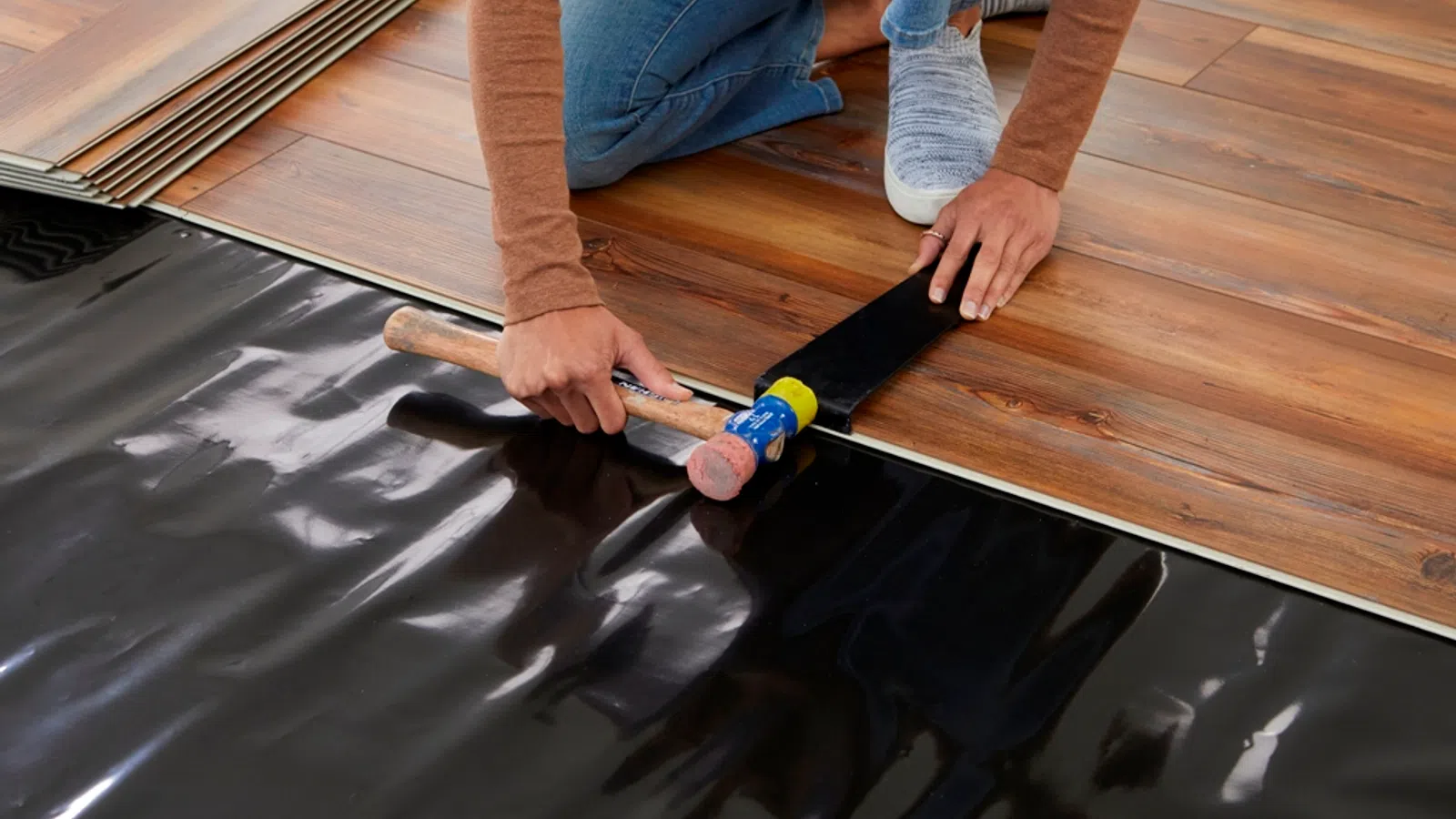 Person installing wood laminate flooring using a rubber mallet and tapping block on black underlayment.