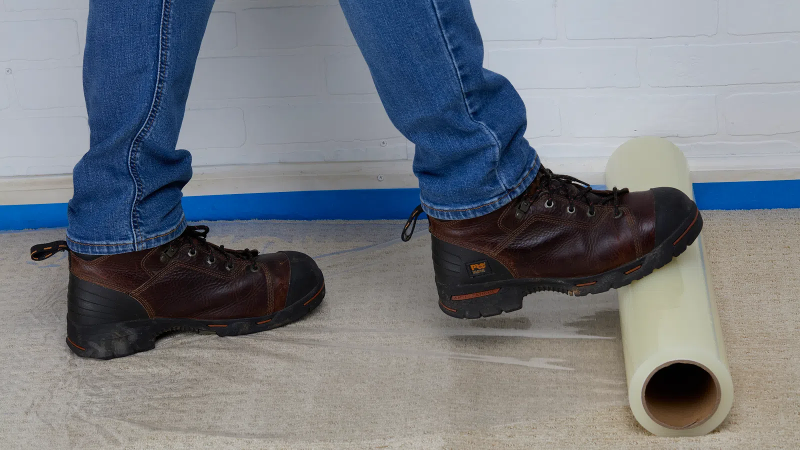 Person wearing brown boots stepping on a beige roll of material on a carpeted floor against a white brick wall.