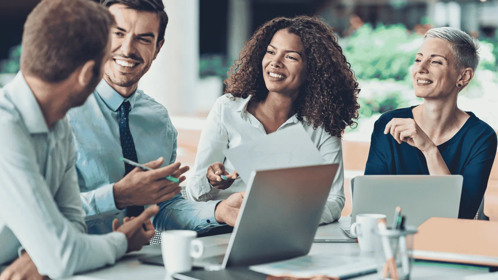 Four diverse professionals smiling and discussing ideas around a table with laptops and documents in a bright office.