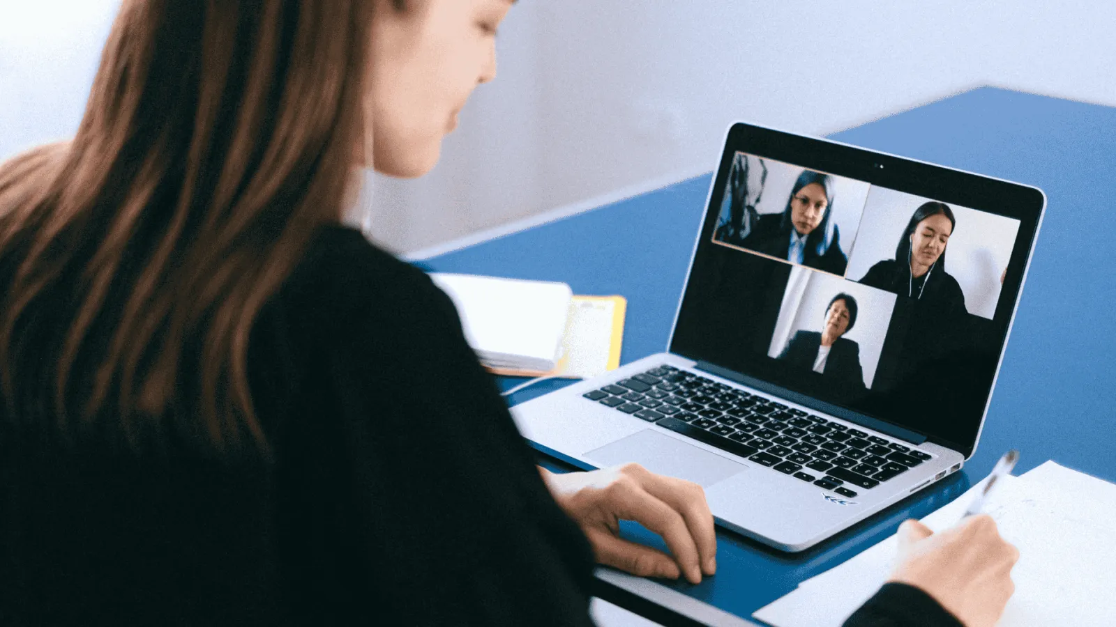 Woman attending online video meeting on laptop, taking notes at a blue desk with notebooks.