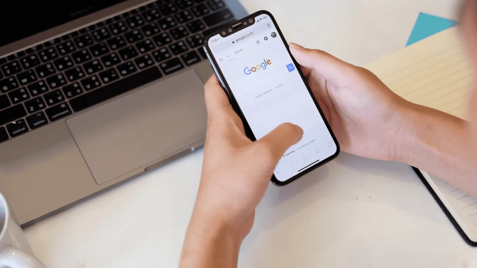 Person holding smartphone with Google search page open next to laptop on desk with notebook.