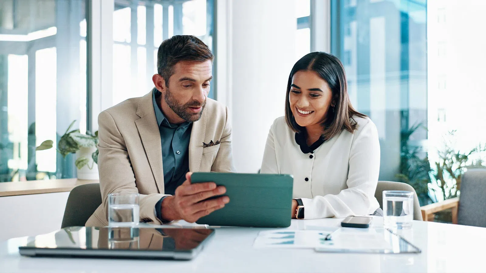 Two professionals collaborating over a tablet in a modern office with large windows and natural light.
