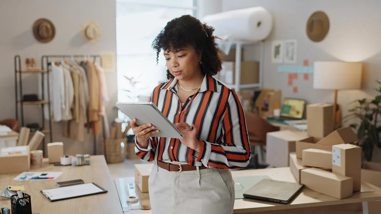 Woman in striped shirt using tablet in a home office surrounded by cardboard boxes and clothing rack.