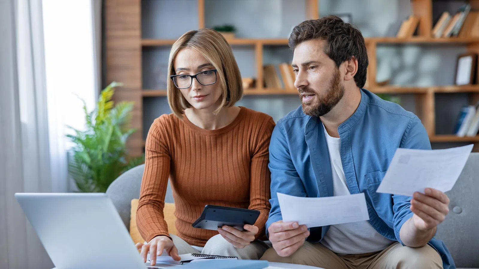 Couple reviewing bills and financial documents together using a laptop at home on a couch.