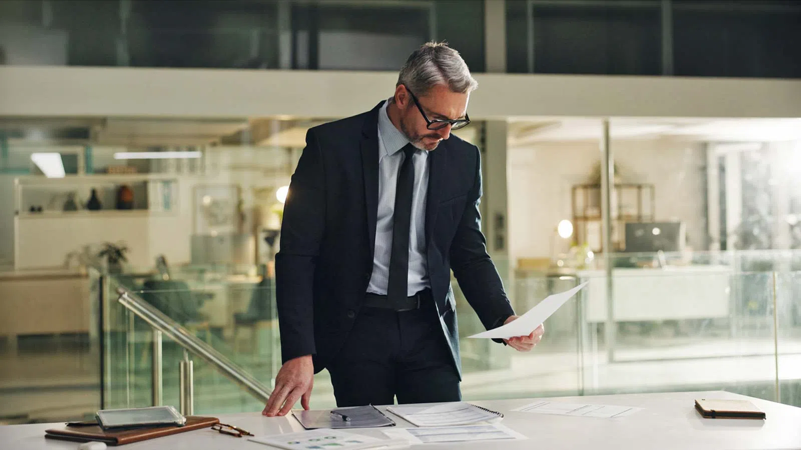 Businessman in black suit reviewing documents at modern office desk with glass walls and office equipment
