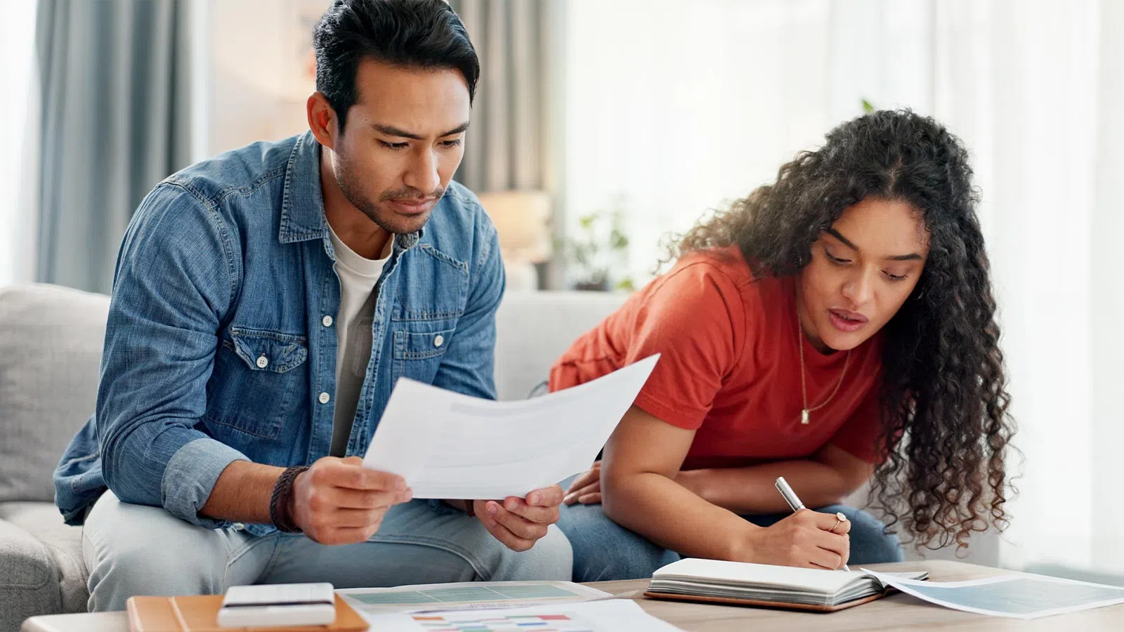 Couple sitting on sofa reviewing documents and taking notes at home with charts and papers on table