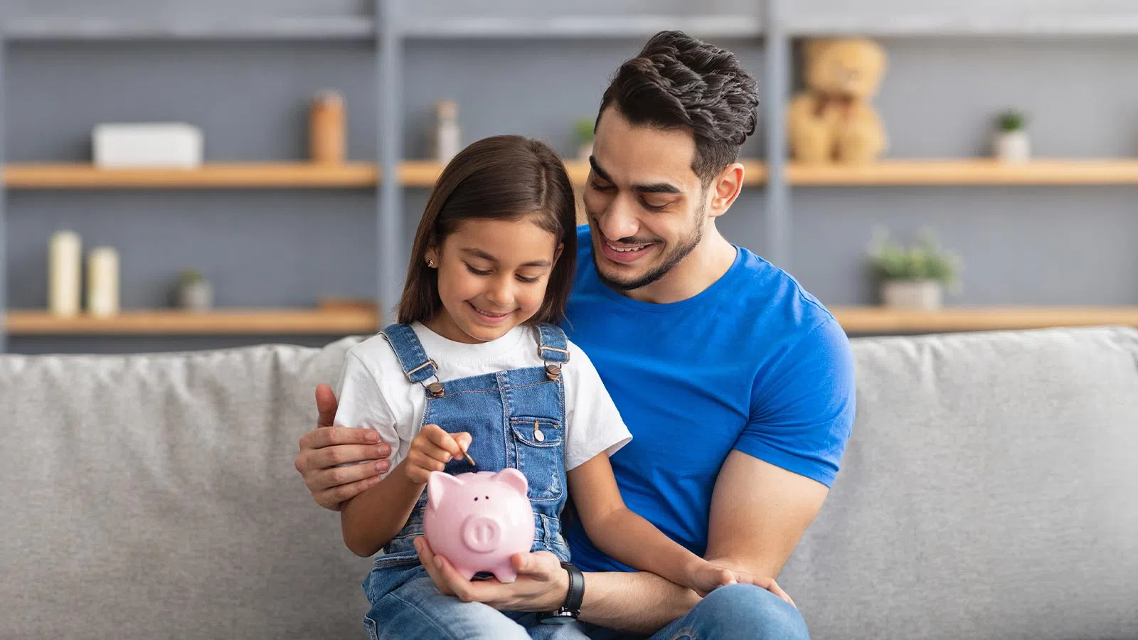 Father and daughter sitting on sofa, happily saving money together using a pink piggy bank