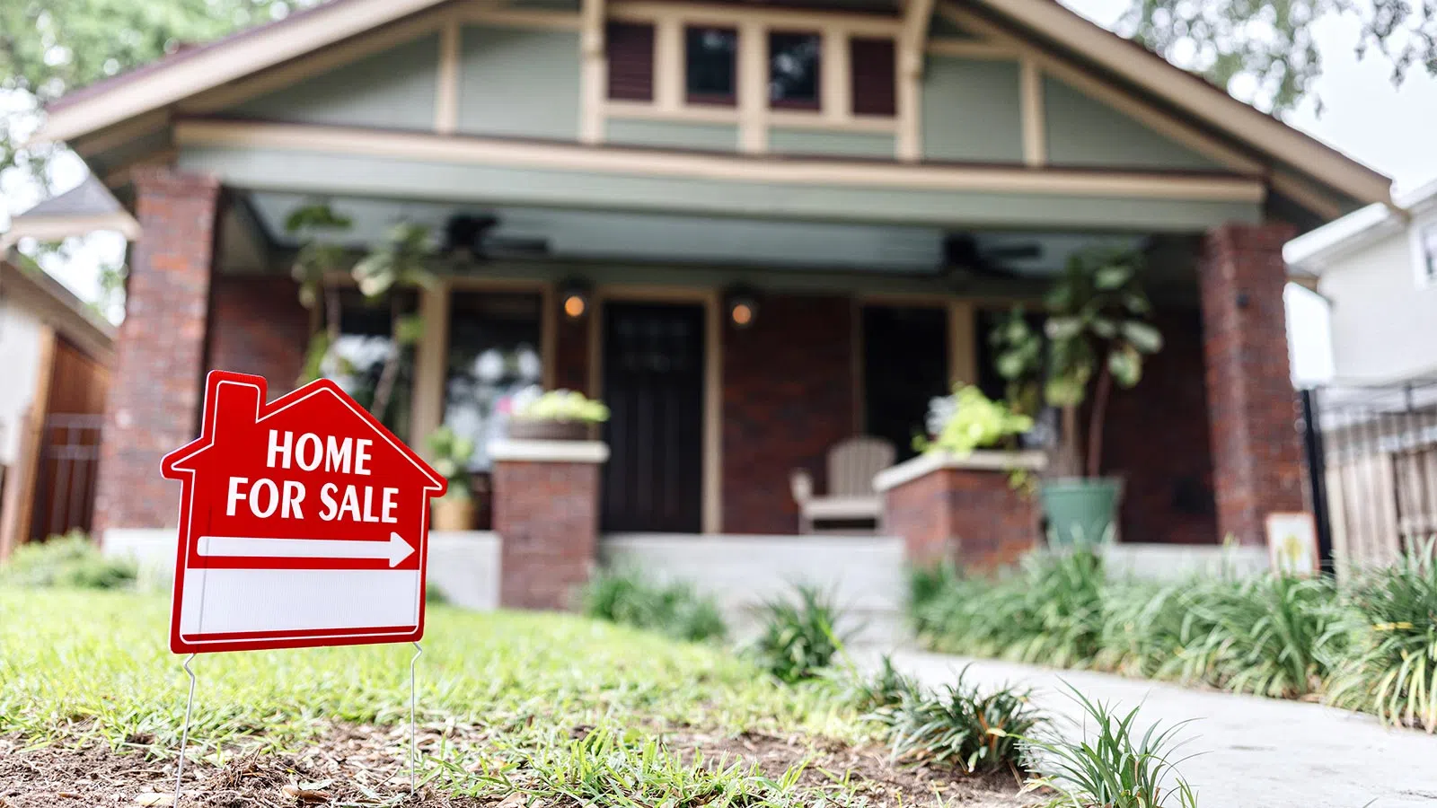 Red Home For Sale sign on front lawn of a brick house with porch and plants in a residential neighborhood.