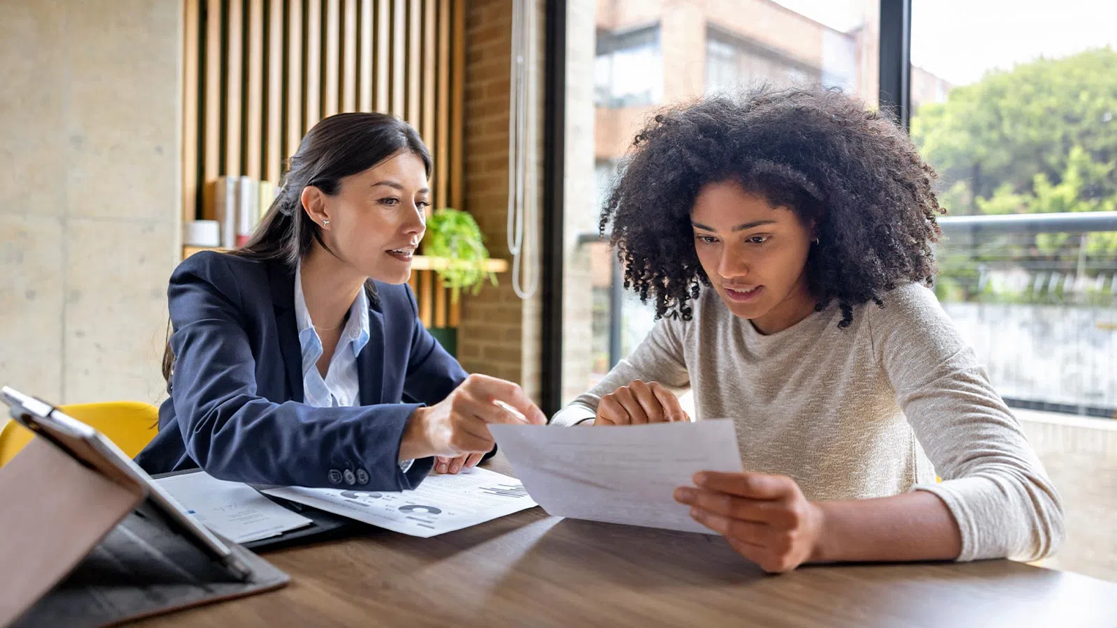 Two professional women reviewing documents together at a table in a bright office with large windows