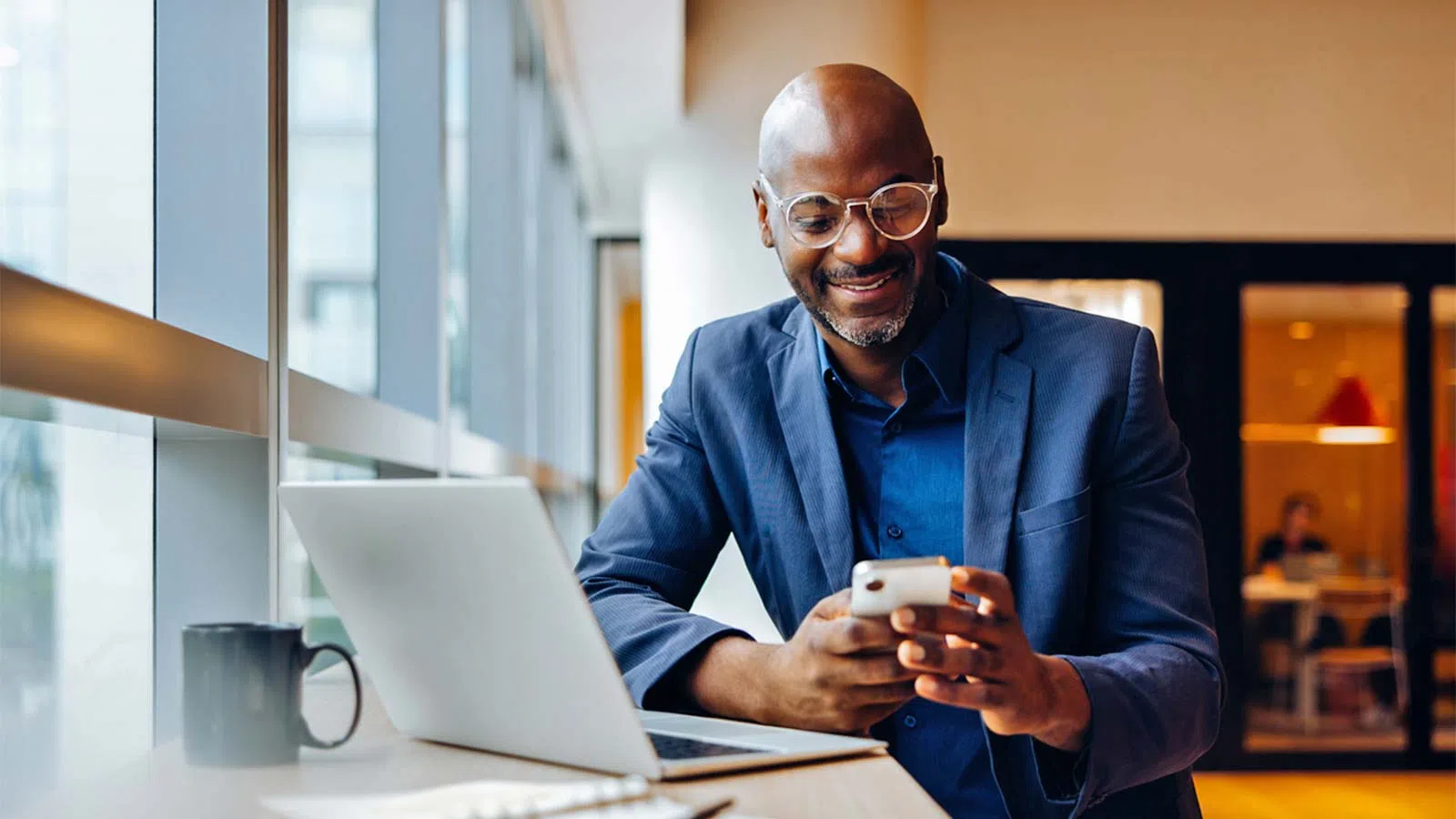 Smiling businessman using smartphone while sitting at a desk with laptop and coffee mug in modern office.