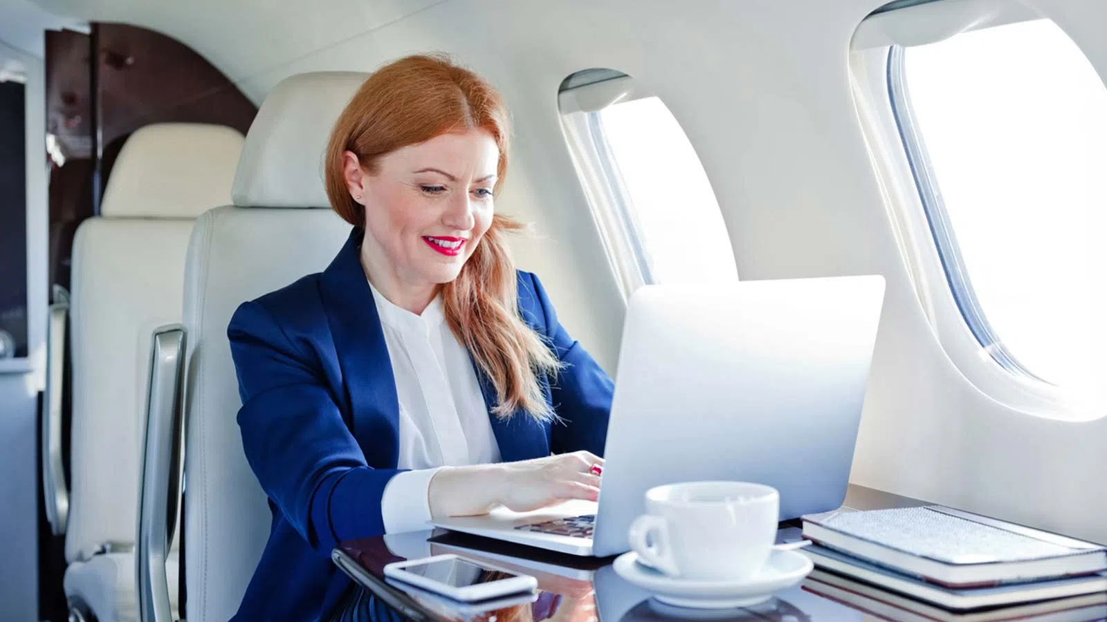 Businesswoman working on laptop inside private jet with coffee cup and notebooks on table by window
