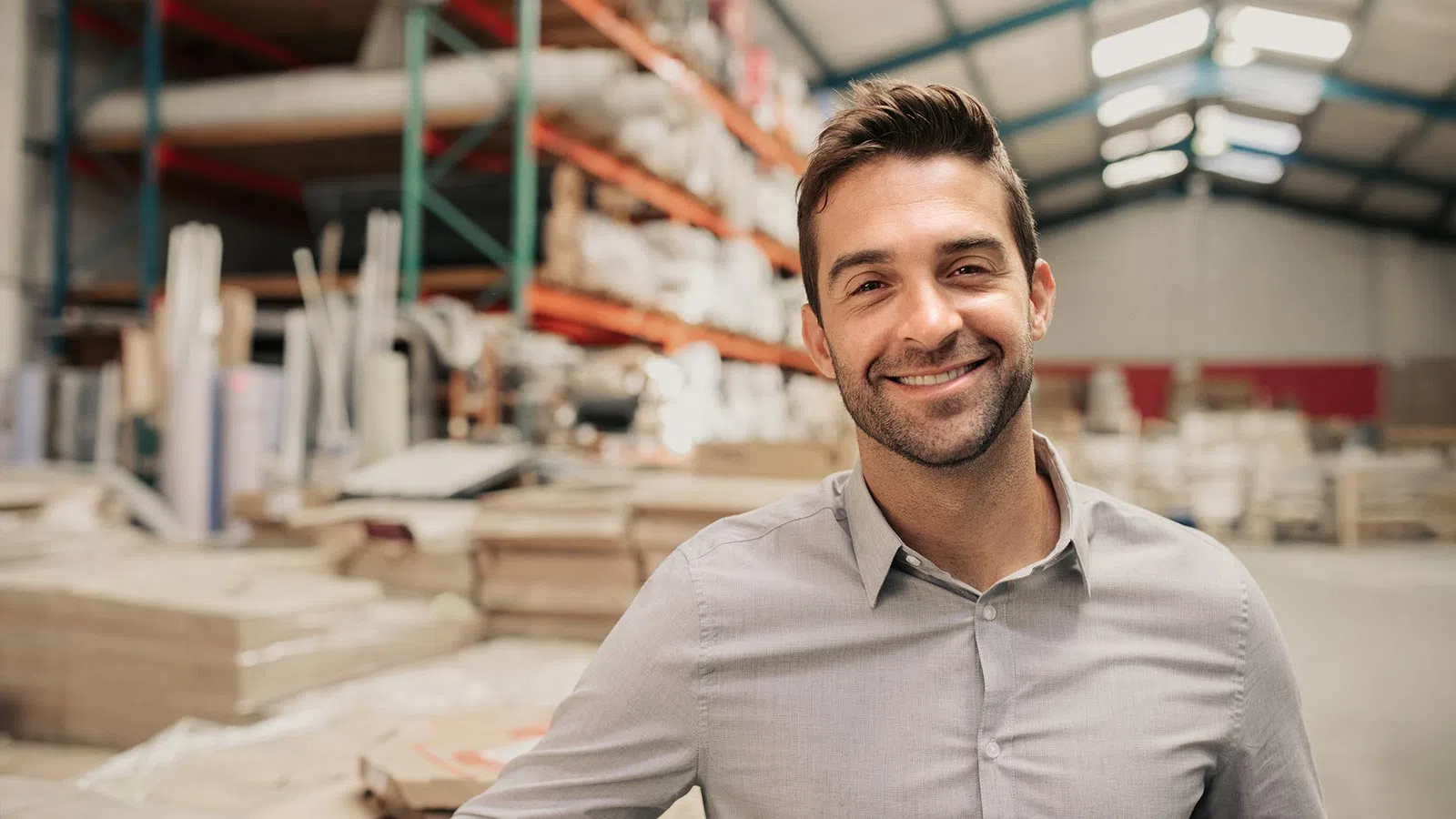 Smiling man in light gray shirt standing confidently in a warehouse with shelves and packaged goods in the background