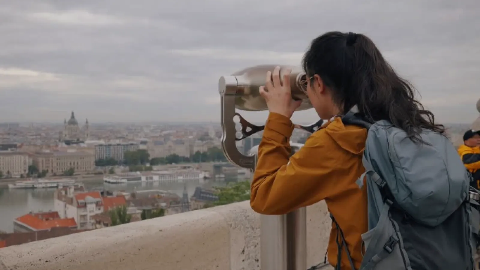 Woman with backpack using a coin-operated binocular viewer overlooking a cityscape with river and historic buildings.