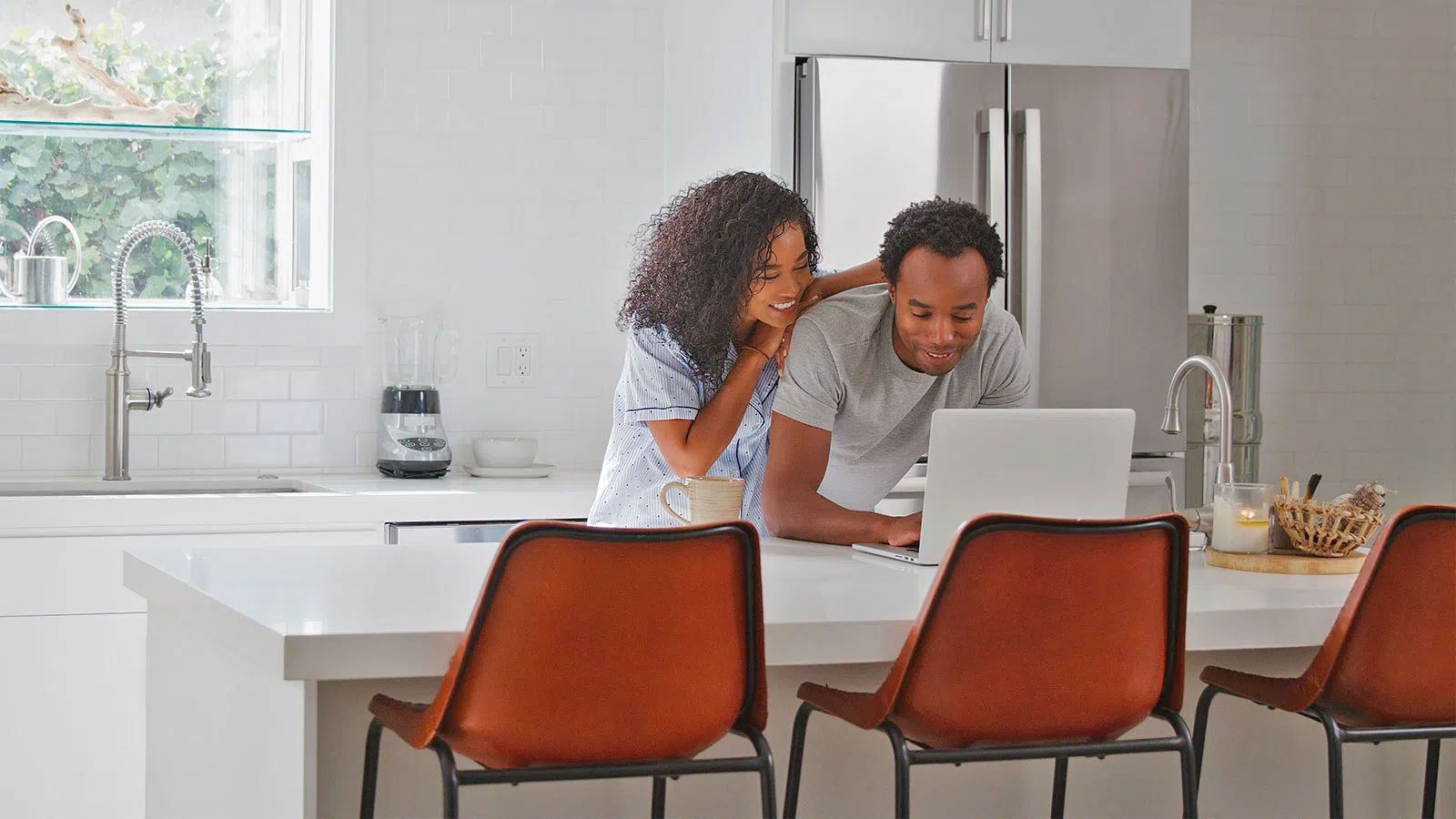 Couple using laptop together while standing at white kitchen island with brown chairs and modern appliances